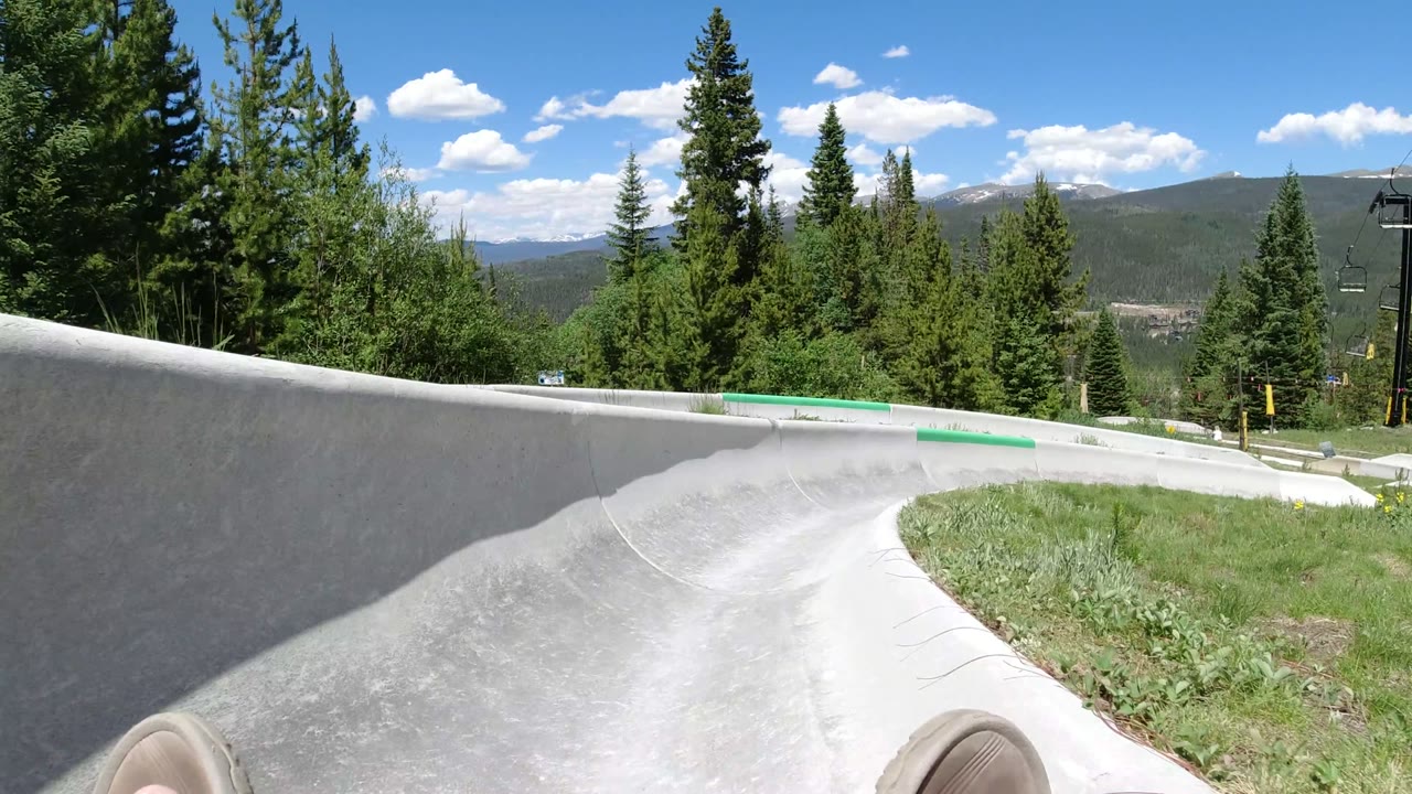 Opening view from a concrete alpine slide in Winter Park, Colorado, with evergreen trees, distant mountains, blue sky, and no visible people.