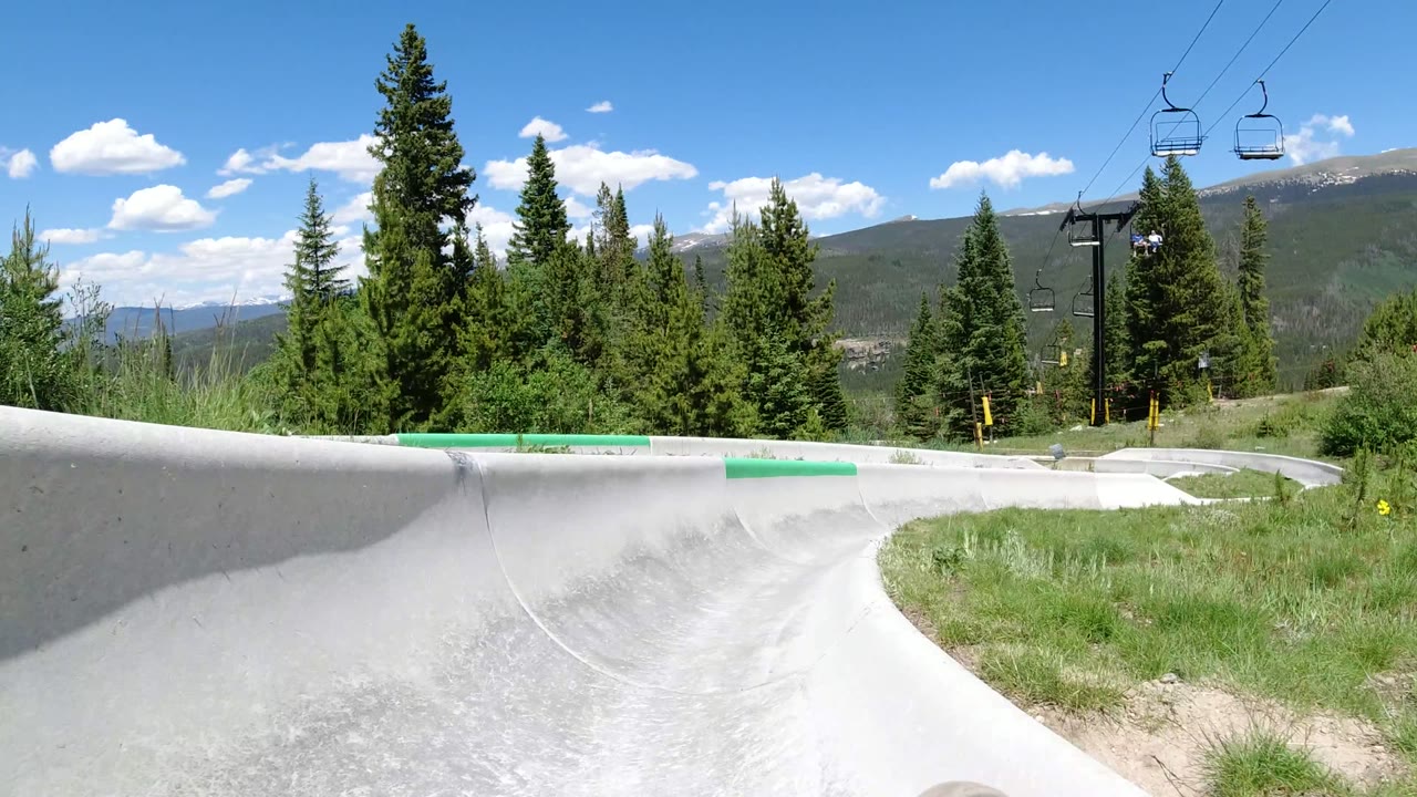 A left-banked section of the concrete slide runs through grass in Winter Park, Colorado, with evergreen trees and mountains behind it.