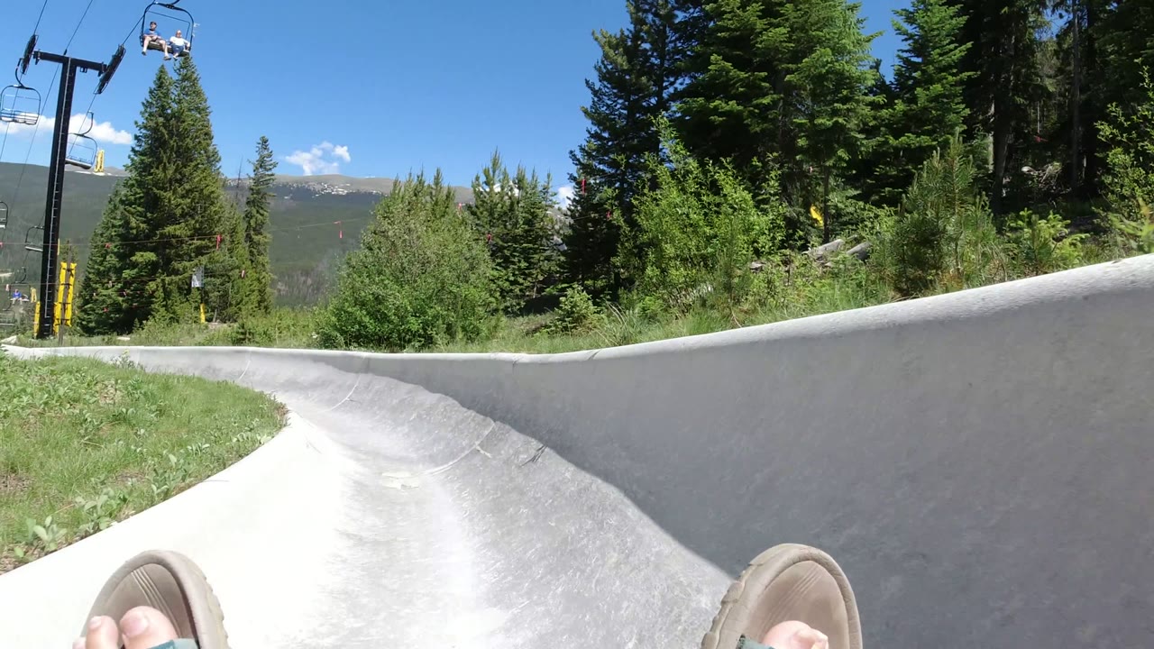 First-person view from a gray outdoor slide in Winter Park, Colorado, with sandal-covered feet at the bottom edge, evergreen trees, chairlift equipment, distant mountains, and bright blue sky.