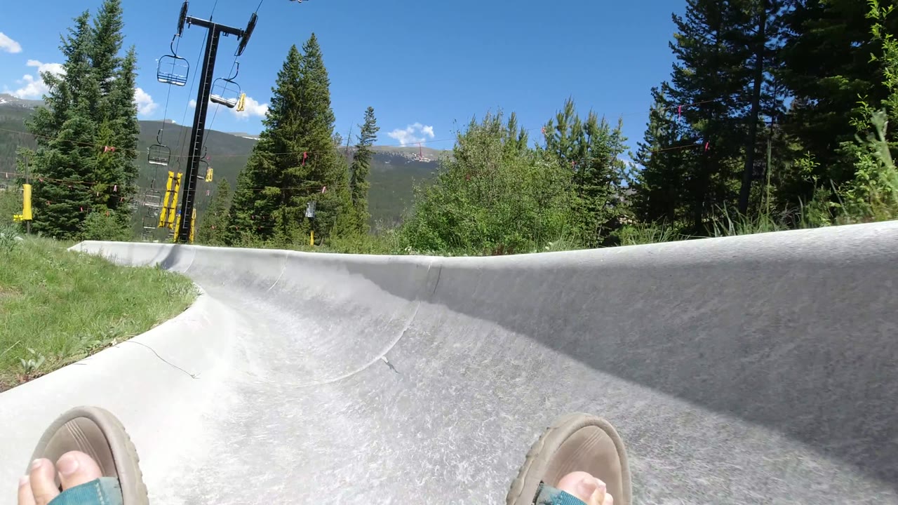 A later POV frame in Winter Park, Colorado shows the slide curving left, with sandals at the lower edge, dense evergreen trees, chairlift poles, distant hills, and clear midday light.