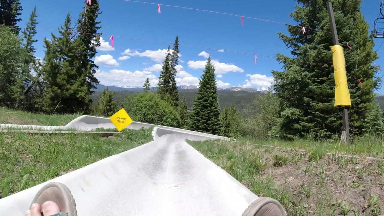 First frame of a Winter Park, Colorado ride POV showing a white chute descending between grass and evergreen trees, with the rider's feet partly visible and no visible faces.