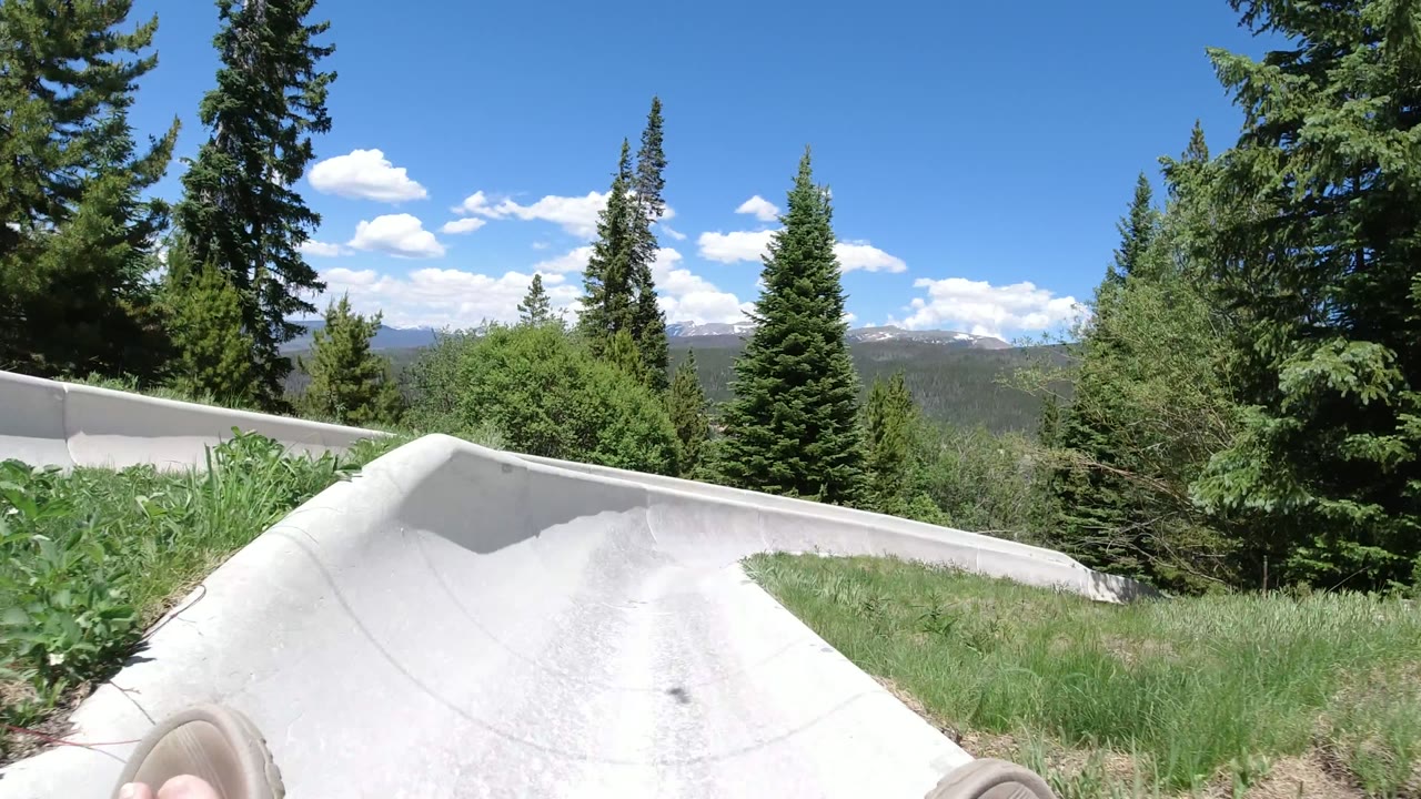 Winter Park, Colorado ride POV looking along a pale curved chute bordered by grass and tall evergreen trees, with the rider's feet at the bottom edge.