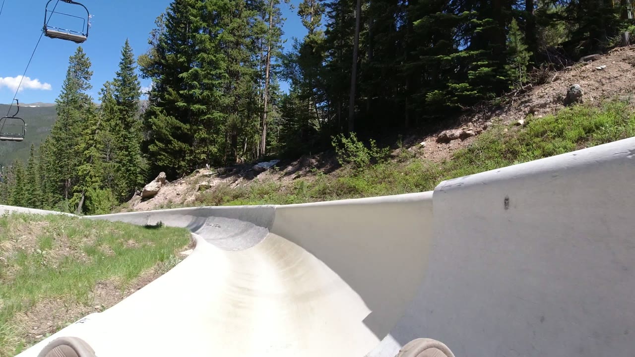 At Winter Park, Colorado, the rider-level view follows a pale concrete slide wall beside a wooded slope, with chairlift chairs overhead and no visible people.