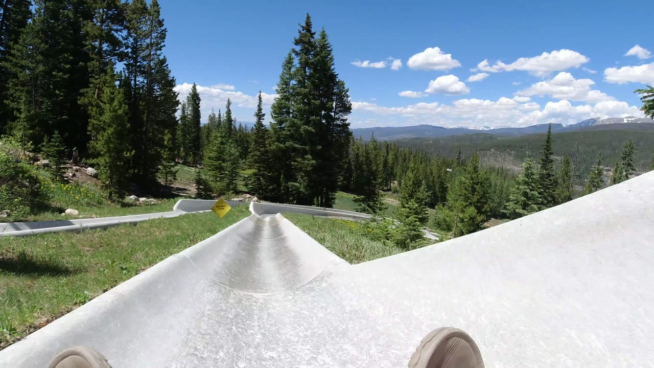 Winter Park, Colorado POV looking down a straight pale slide track bordered by grass and evergreen trees, with forested hills on the horizon and partial rider feet visible at the lower corners.