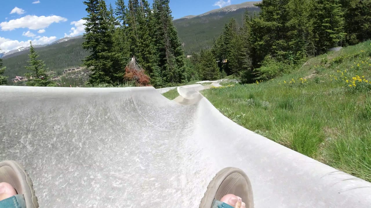 The slide bends through tall green grass in Winter Park, Colorado, with the rider's feet partly visible and forested mountain slopes under bright sun.