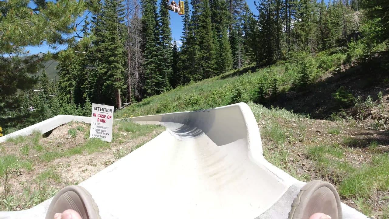 First-person view in Winter Park, Colorado at the start of a pale slide pipe, with slippered feet at the bottom edge, grass and evergreens around the track, chairlift cables overhead, and a warning sign on the left.