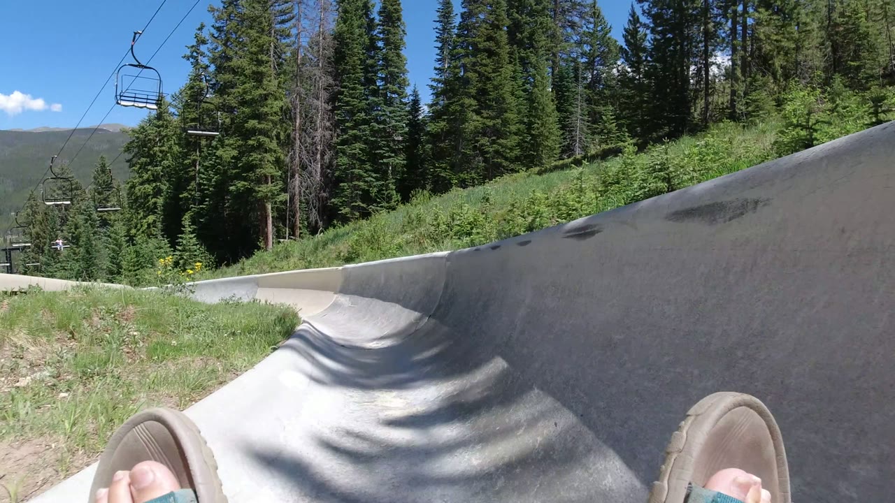 A low rider POV in Winter Park, Colorado shows the pipe wall rising on the right, slippered feet in the foreground, dense evergreen trees beyond, and chairlift chairs hanging above the route.
