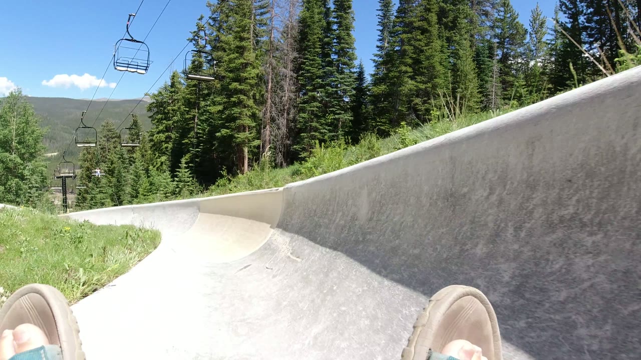 In Winter Park, Colorado, the pipe wall fills the right side of the frame while slippered feet appear at the bottom, with grass, evergreen trees, chairlift chairs, and a distant ridgeline visible beyond the rim.