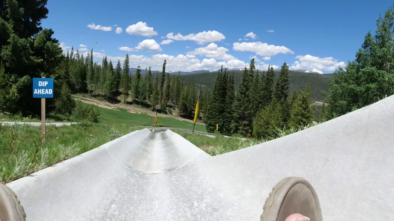 The Winter Park, Colorado slide curves downhill past grass and trees, with a blue dip ahead sign beside the concrete track and the rider's sandal partly visible.