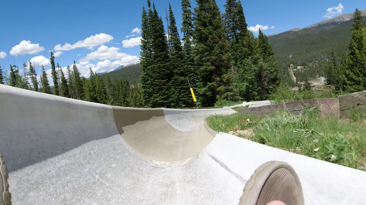 Feet at the front of a pale slide track with evergreen trees and mountains behind.