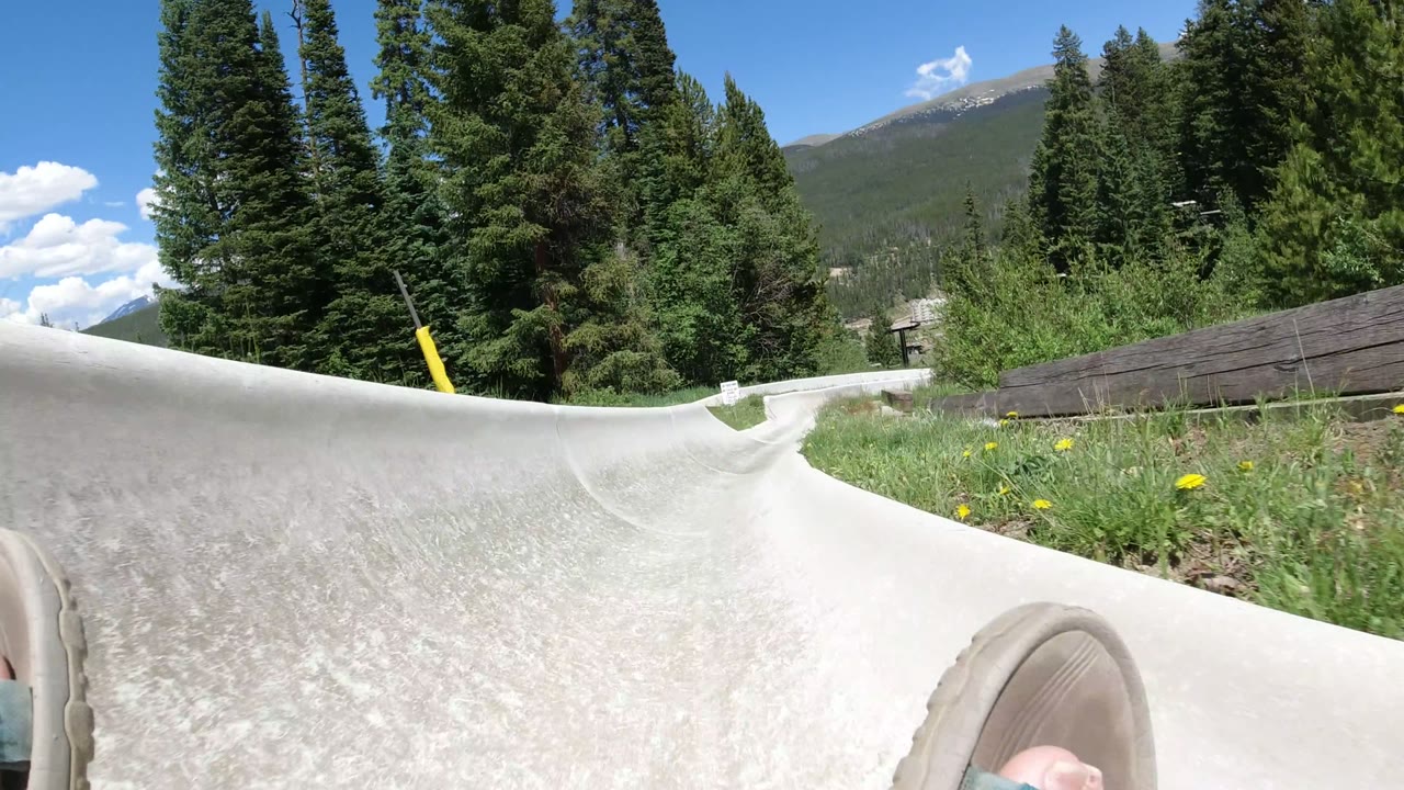 A Winter Park, Colorado POV frame looks down a light concrete slide curve, with the rider's sandal at the bottom edge, dense evergreens, grasses, and a mountain ridge beyond.