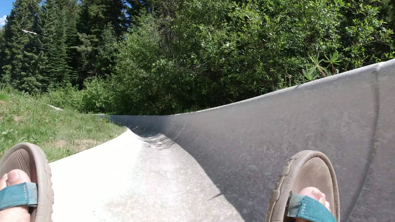 The rider's sandals stay in the foreground as the stone slide bends past tall grass and dense green trees in Winter Park, Colorado.