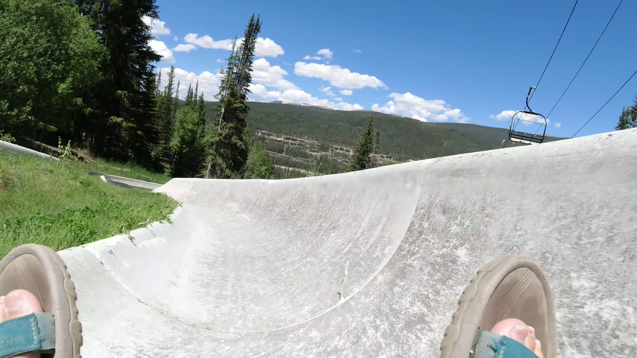 First-person view in Winter Park, Colorado with sandaled feet at the bottom of the frame, a pale concrete luge track ahead, evergreens, chairlift cables, and blue sky.