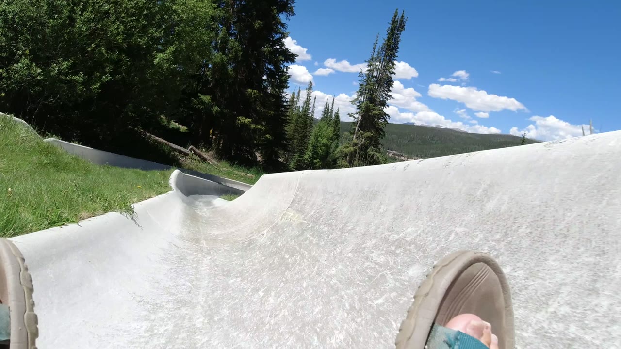 The POV continues along the Winter Park, Colorado luge track, with one sandal at the lower edge, a banked concrete wall, grass, evergreens, and distant hills.