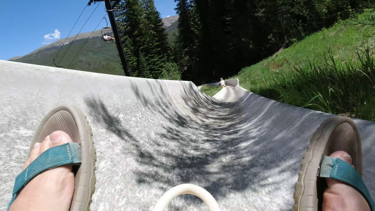 First-person view in Winter Park, Colorado with sandal-clad feet entering a pale slide, evergreen trees at the sides, lift cables overhead, and mountains under bright sun.