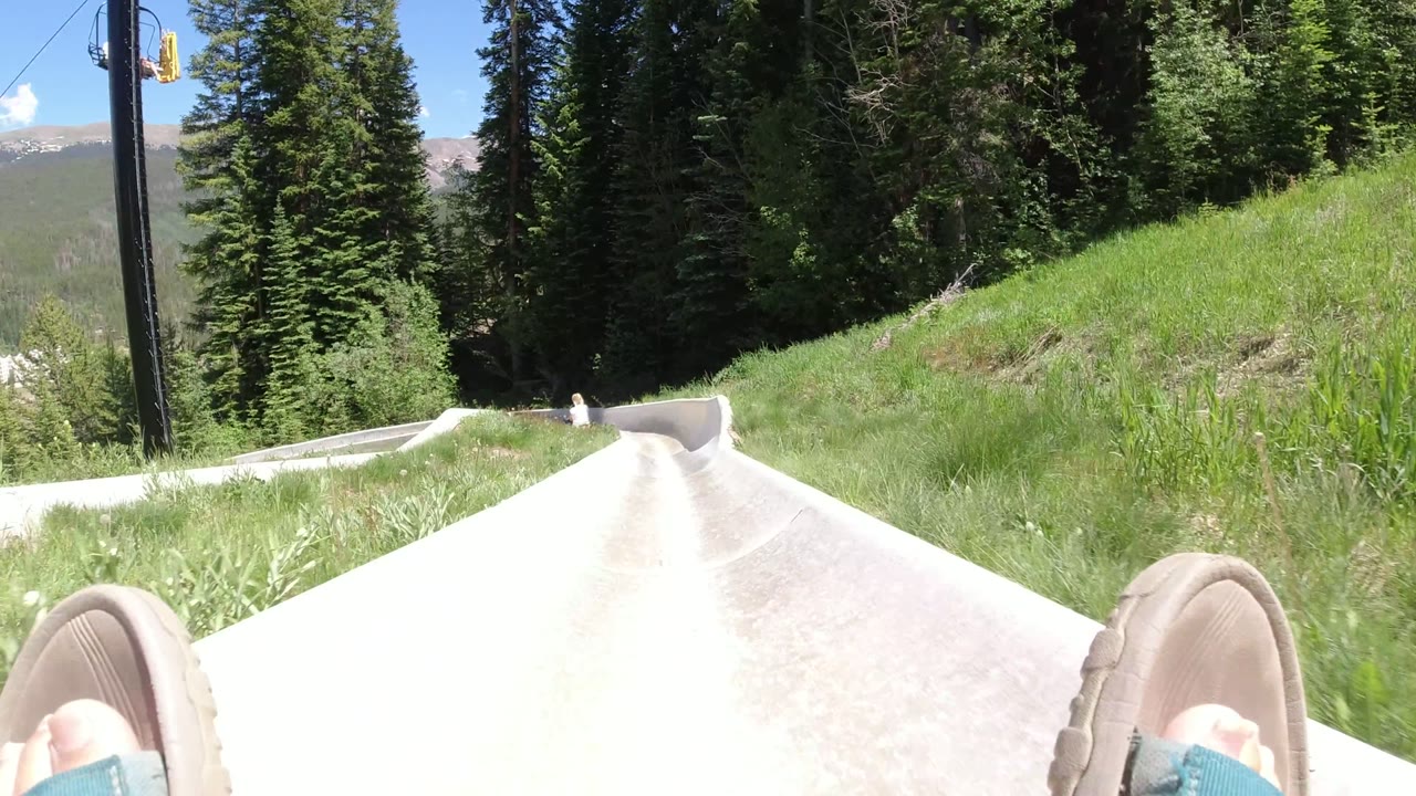 First-person Winter Park, Colorado frame looking down a pale slide channel, with sandal toes in the foreground, green grass on the banks, and dense evergreens ahead.