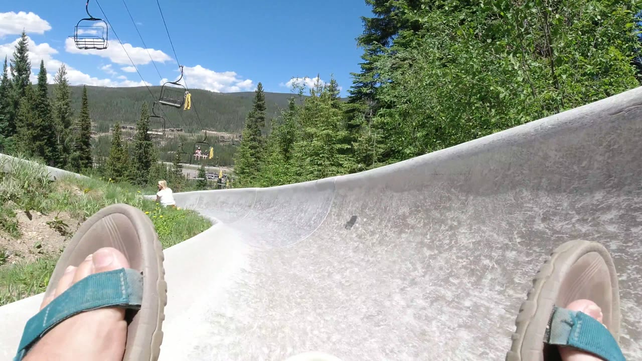 POV on a gray slide with sandaled feet, trees, chairlift seats, and mountains.