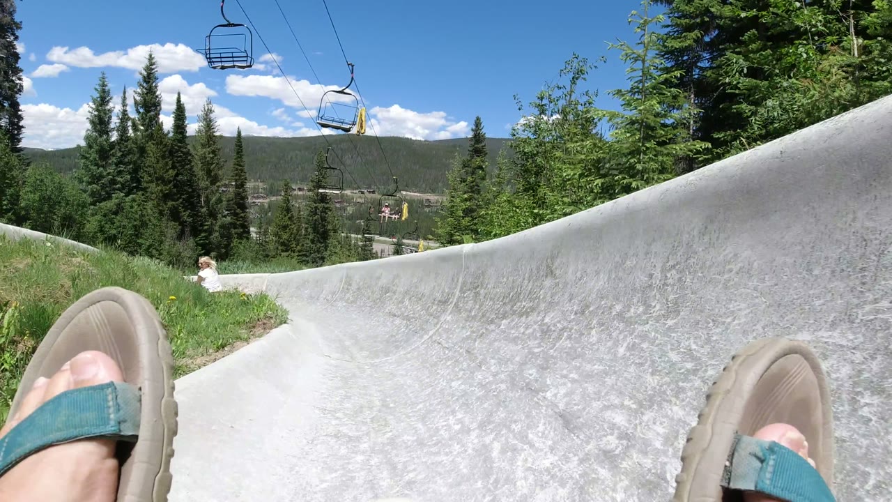 At 0:01 in Winter Park, Colorado, the slide curves past grass and trees with sandaled feet in the foreground and a small child visible farther down the track.
