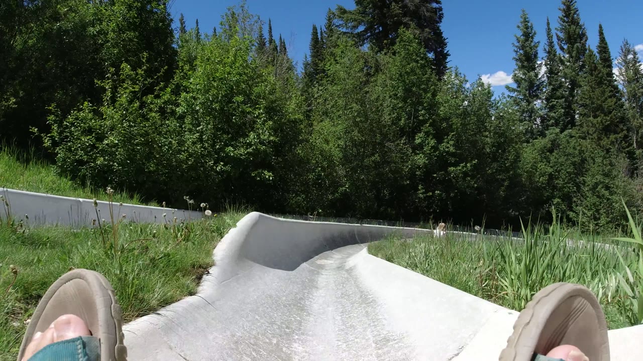 First frame of a Winter Park, Colorado concrete attraction slide, with the rider's sandaled feet at the bottom edge, grass on both sides, and dense trees ahead.
