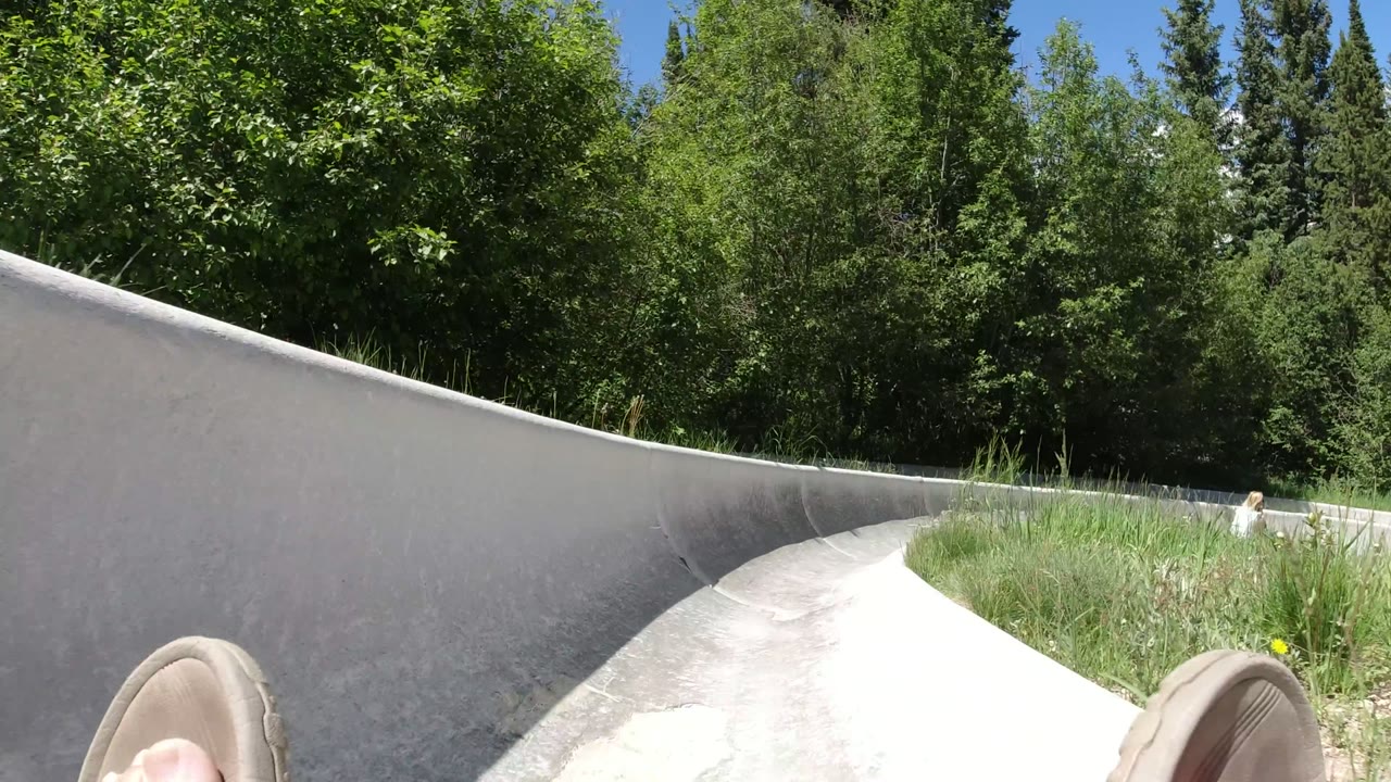 A low first-person view inside the Winter Park, Colorado slide shows the gray concrete wall rising on the left, the rider's sandal at the bottom, and leafy trees filling the background.