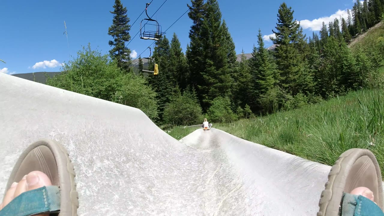 Rider POV on a white alpine slide track in Winter Park, Colorado, with sandal-clad feet at the bottom edge, green trees ahead, and chairlift hardware above.