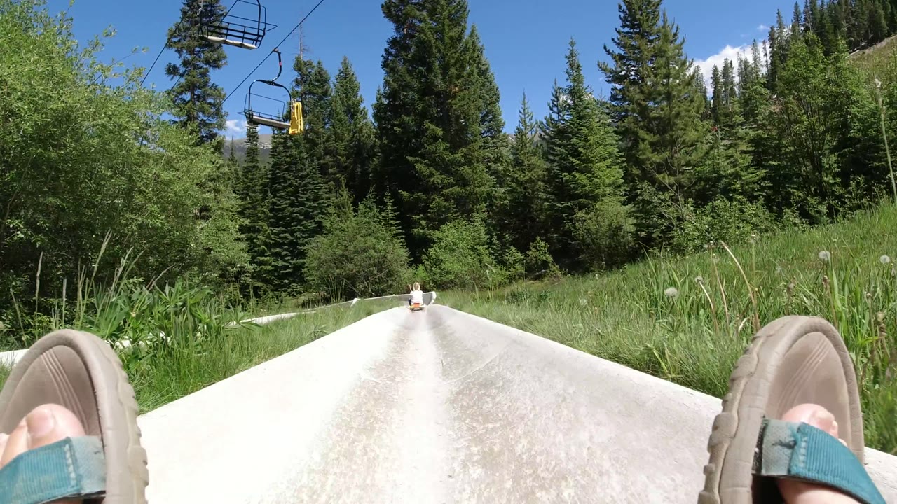 The Winter Park alpine slide track narrows into the distance between tall conifers, with the rider's feet low in frame and bright midday sky above.