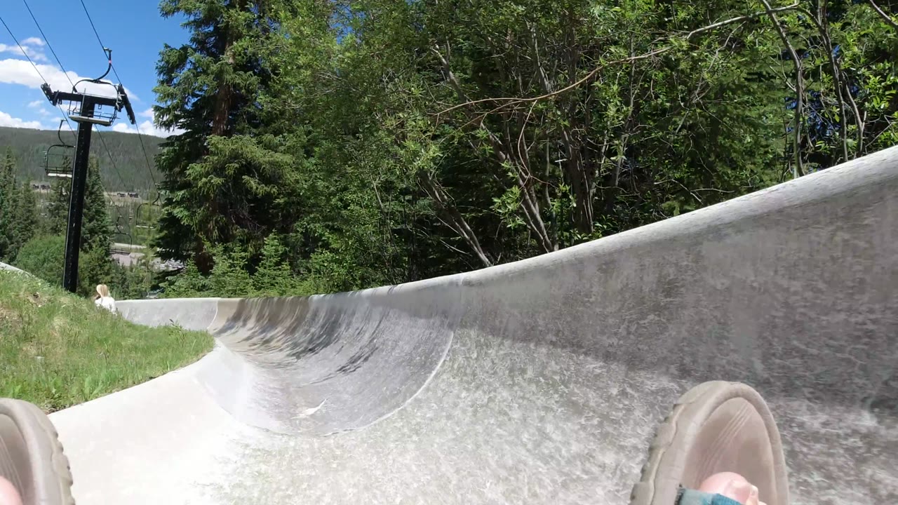 Curving concrete slide with sandaled feet, trees, chairlift cables, and blue sky.