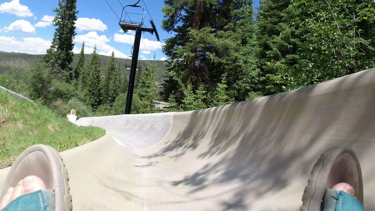 Winter Park, Colorado alpine slide curving through grass and trees, with the rider's feet partly visible, a chairlift tower above the channel, and no recognizable people.