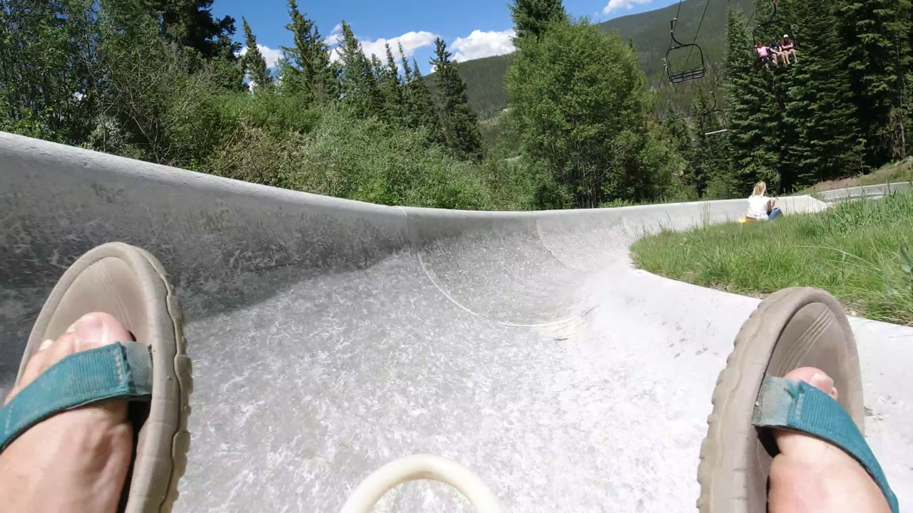 First frame of a Winter Park, Colorado slide ride, with teal sandals in the foreground, a gray slide curving ahead, evergreen trees, blue sky, and chairlift riders in the distance.