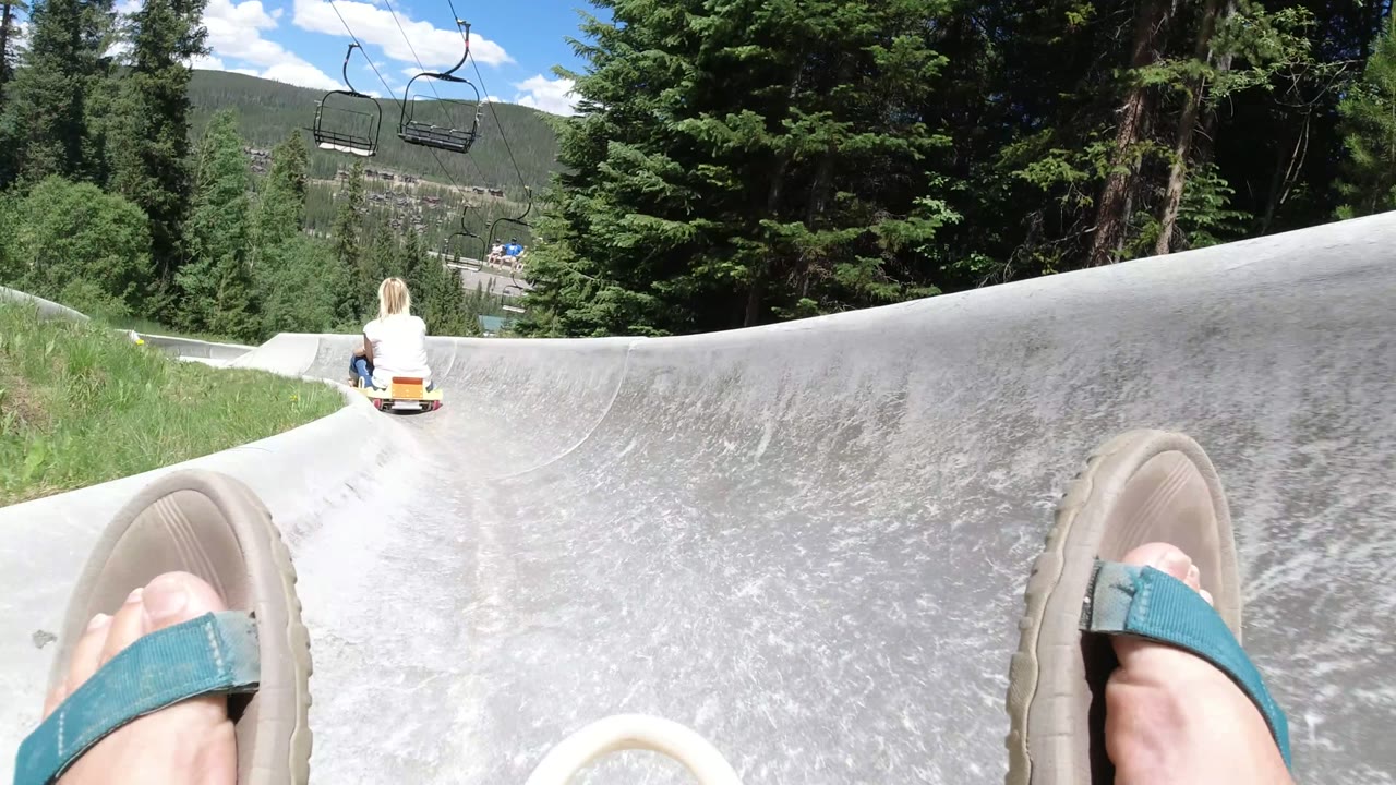Feet in sandals on a concrete slide behind a cart rider in a forested mountain setting.