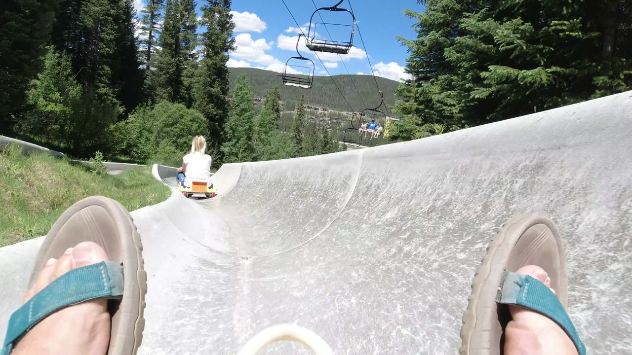 Winter Park, Colorado frame showing the concrete slide banking left, a cart rider ahead, sandals in the foreground, dense evergreens, grassy edges, and suspended chairlift seats.