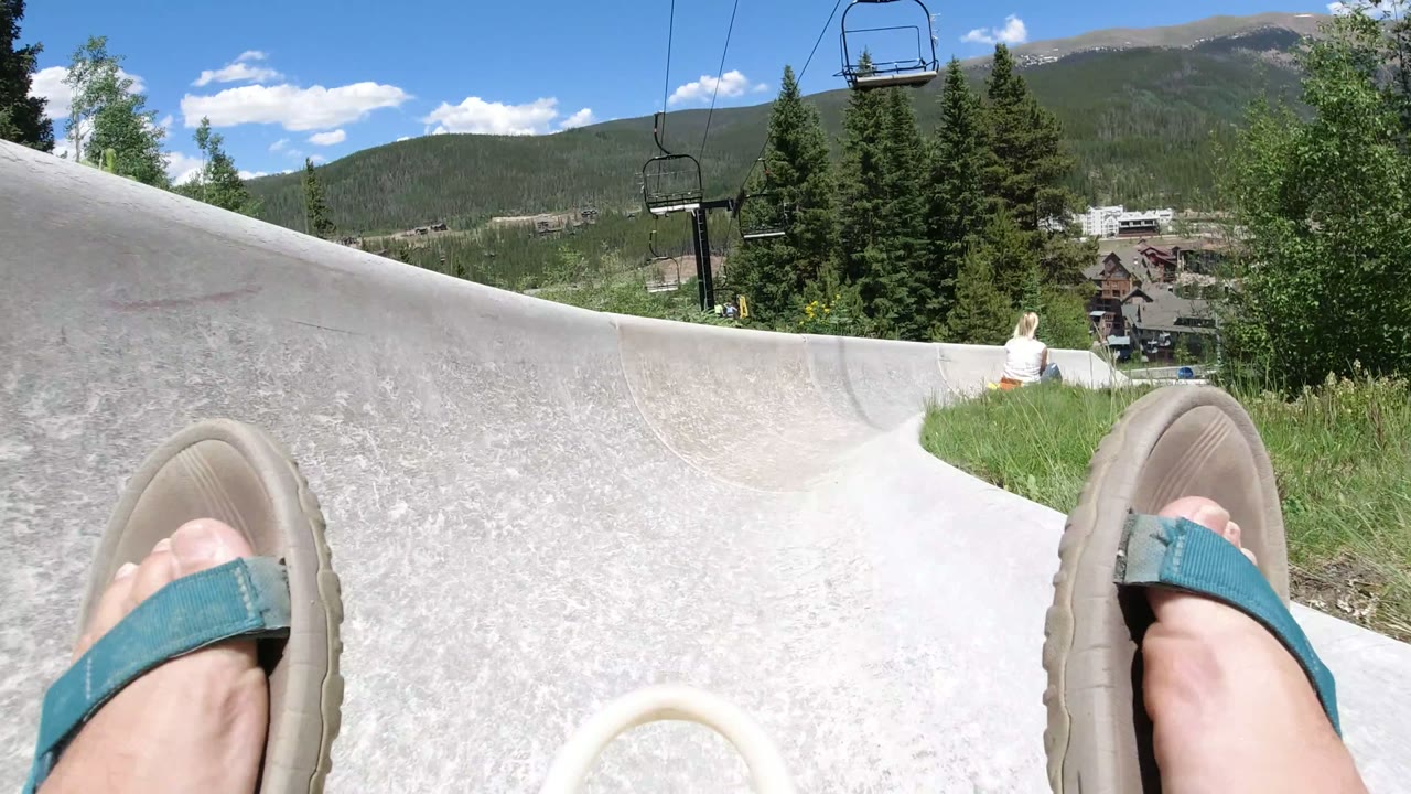Sandaled feet in an alpine slide chute above trees and mountains.