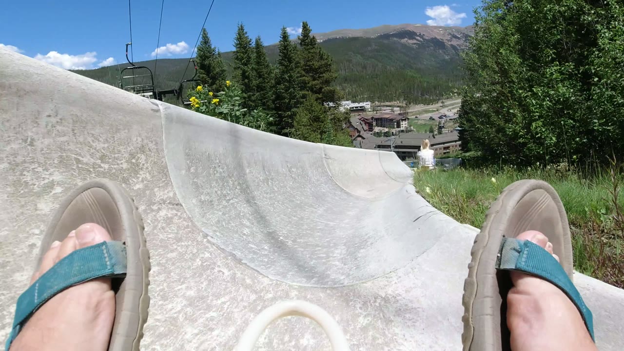 Winter Park, Colorado slide POV looking into a curving concrete chute, with the rider's sandals low in frame, chairlift equipment overhead, evergreens, resort buildings, and mountains beyond.