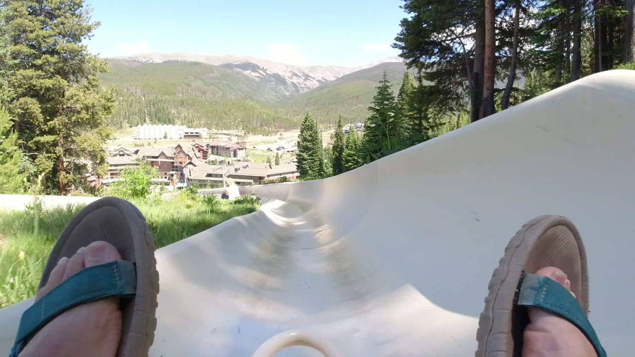 Sandal-clad feet at the start of a pale outdoor slide with trees and mountains beyond.