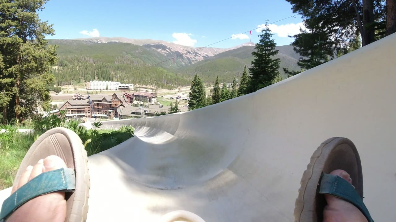 At one second, the Winter Park, Colorado view widens across the slide curve, with one sandal near the left edge, another at right, and distant buildings framed by trees and mountains.