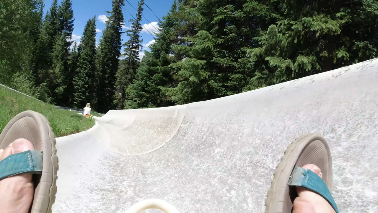 Feet in teal sandals on a pale slide track surrounded by evergreens.