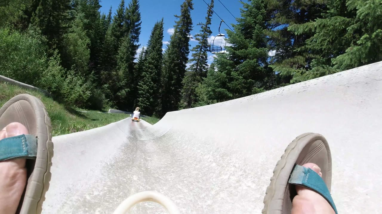 Winter Park, Colorado slide track curving through green grass and evergreens, with the rider's sandals large in the foreground and a chairlift chair above.