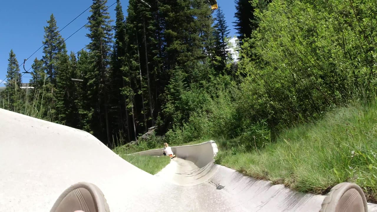 First frame of a Winter Park, Colorado ride POV with sandaled feet in the foreground, a pale curved chute, tall grass, evergreen trees, and no visible faces.