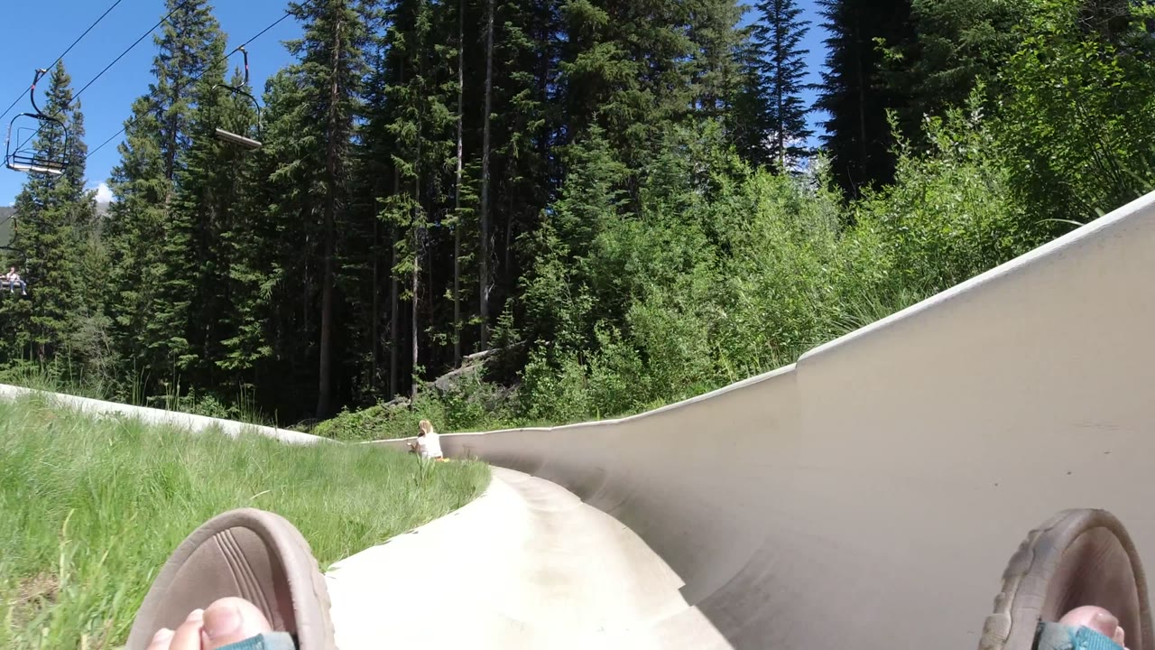 Winter Park, Colorado POV looking down a pale curved ride chute, with tall grass on the inside bend, evergreen forest behind it, and a small distant rider ahead.