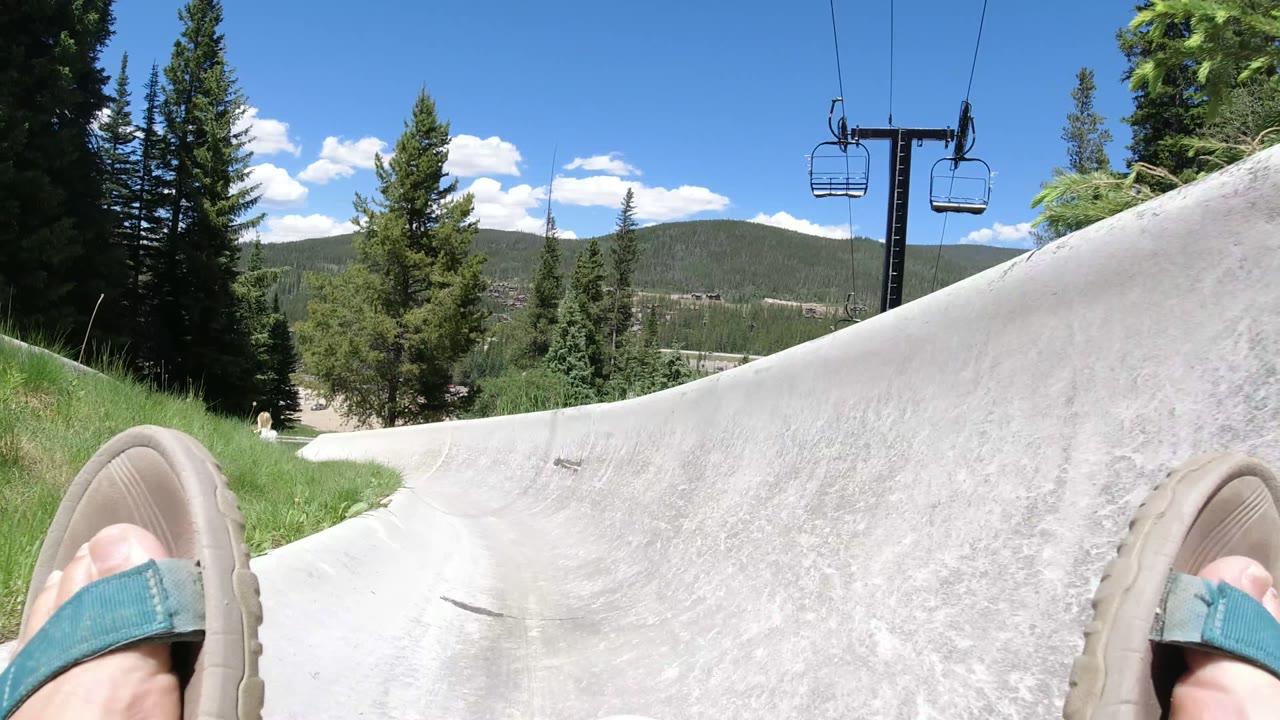 Winter Park, Colorado frame showing the rider's sandaled feet closer to the track edge, a banked concrete curve, green grass, chairlift chairs overhead, evergreens, and distant mountains.