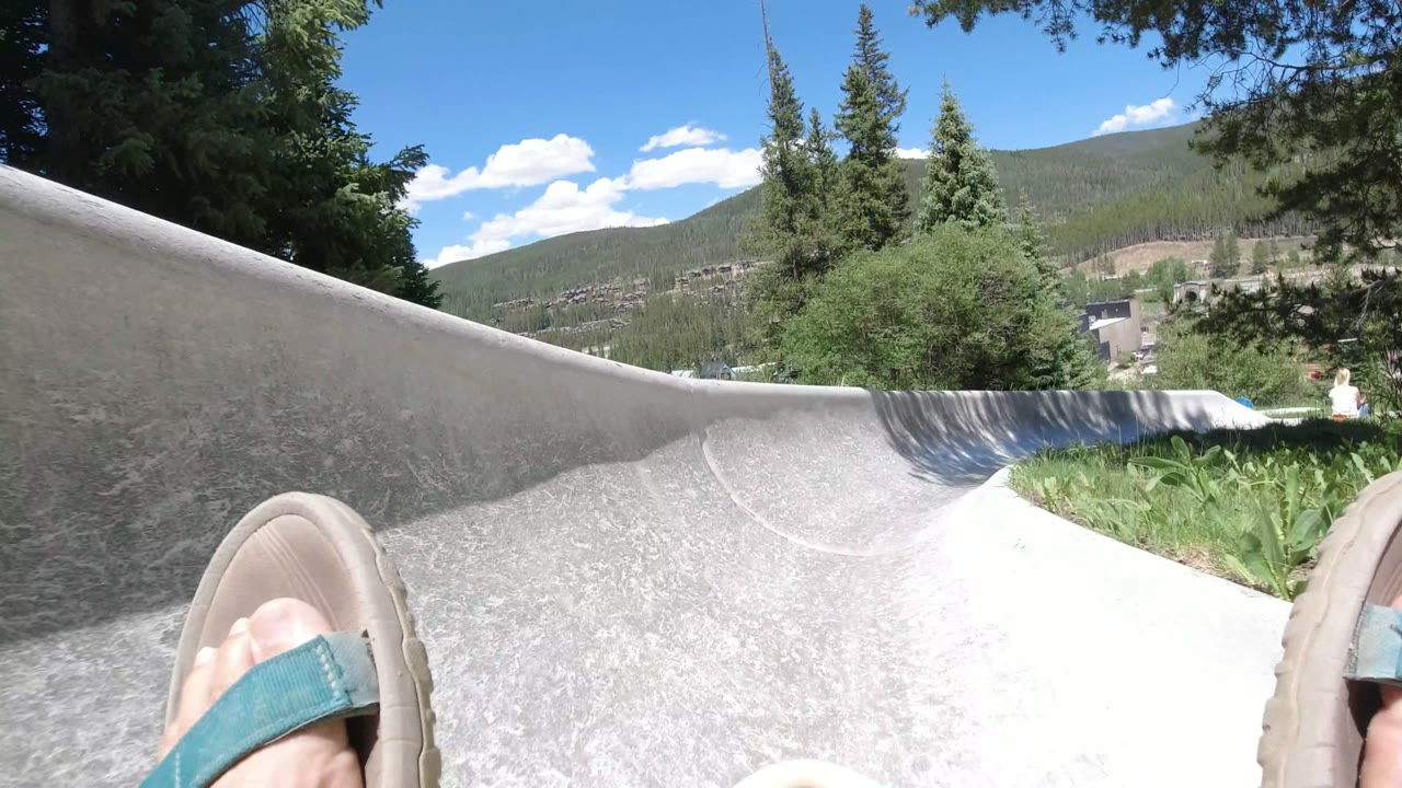 A low POV frame in Winter Park, Colorado shows the rider's sandal near center, the concrete slide rising at left, shrubs and pines at right, and mountain slopes under bright sky.