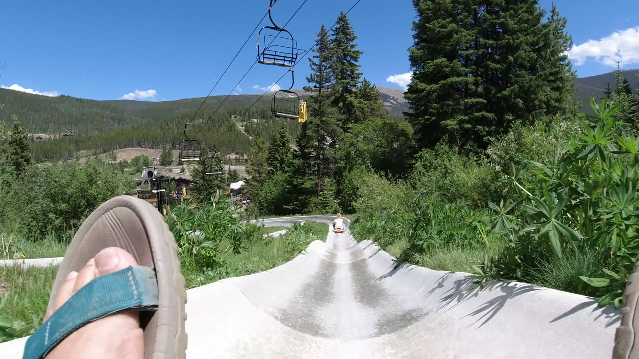 Feet in sandals point down a concrete slide with chairlift seats and mountains beyond.