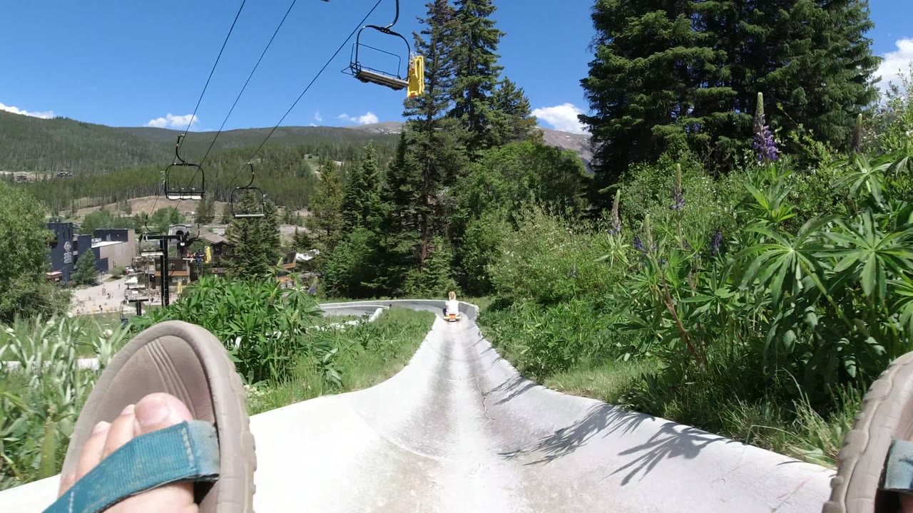 A later Winter Park, Colorado frame keeps the feet low in view while the concrete chute narrows toward shrubs, pine trees, overhead lift chairs, distant structures, and a forested mountain horizon.