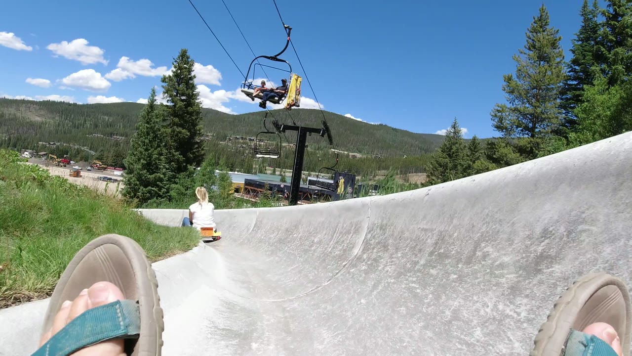 Rider POV on a concrete alpine slide with a child ahead and chairlift overhead.