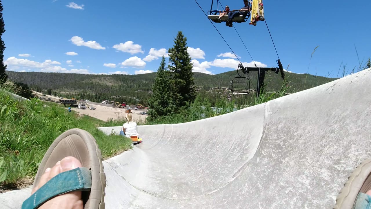 Winter Park, Colorado frame shows the slide channel rising along the right edge, the rider's sandal in the foreground, a blonde child on the track, trees, chairlift equipment, and distant mountains.