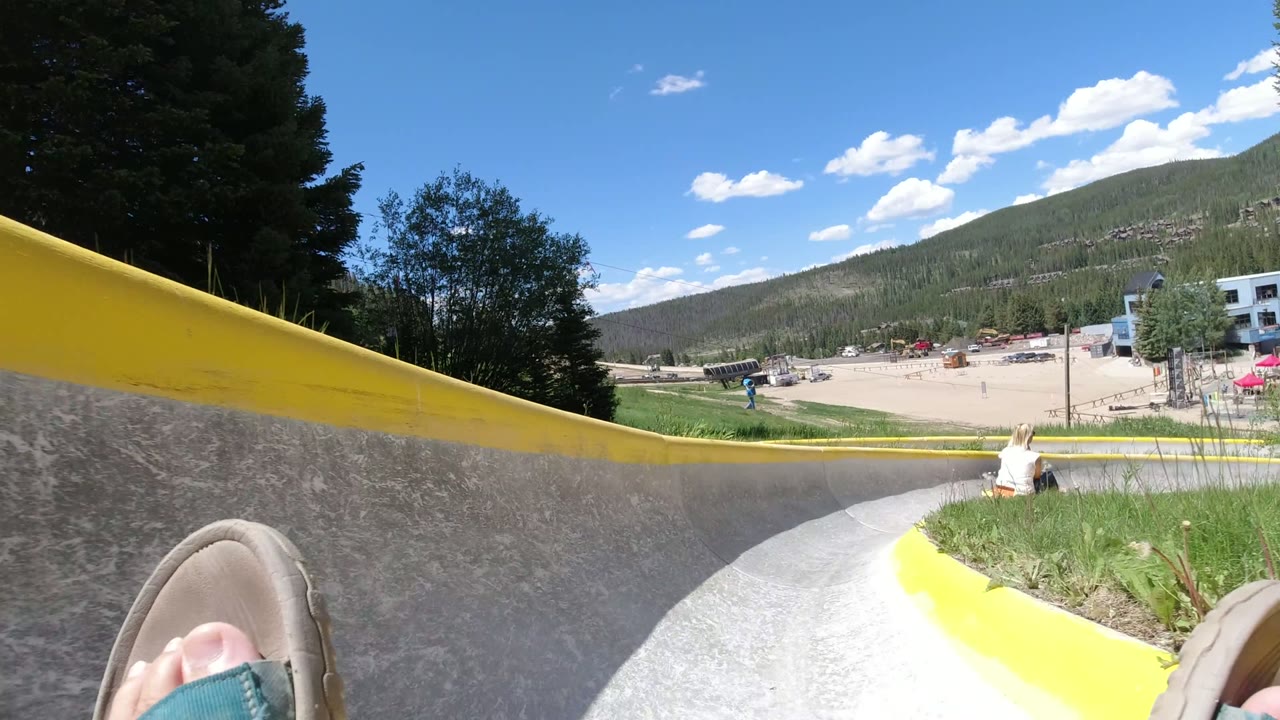 POV from an alpine slide track with rider feet, yellow rails, trees, buildings, and mountains.