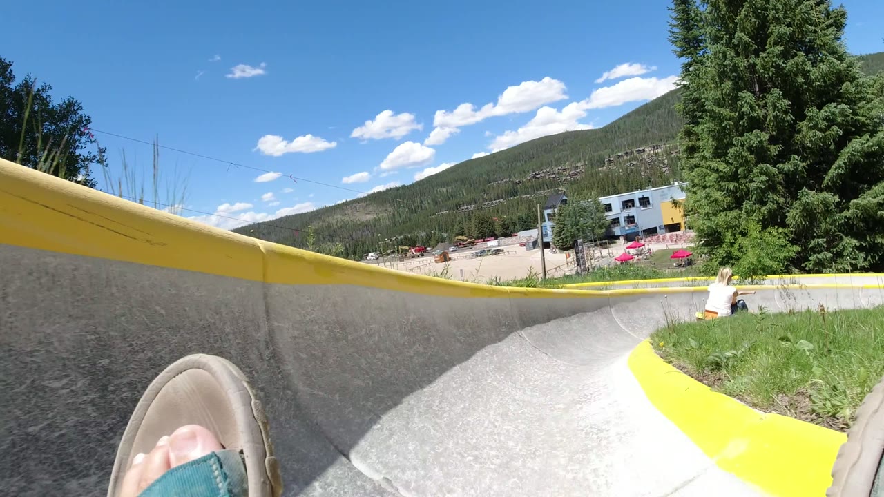 Winter Park, Colorado alpine slide POV rounding a curve, with the rider's shoes in the foreground, yellow rails, a small rider ahead, trees, buildings, and a mountain ridge.