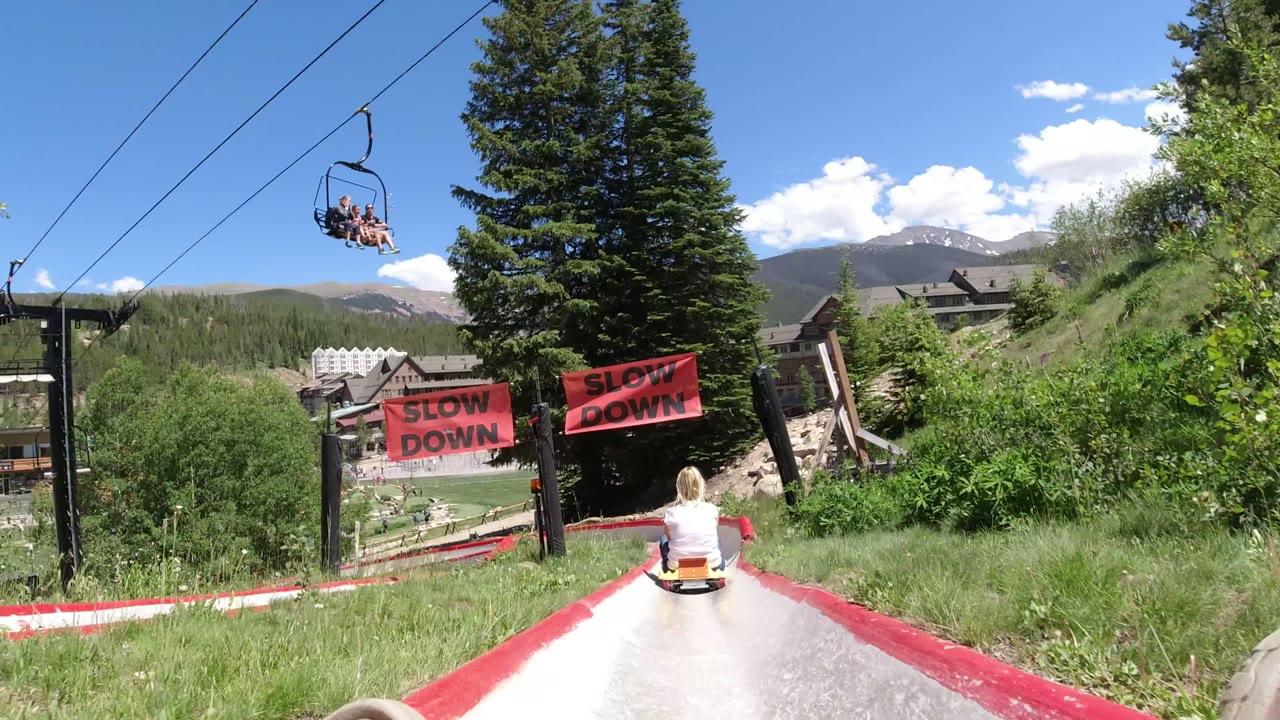 At Winter Park, Colorado, a rider heads down a concrete alpine slide with red rails, chairlift cables overhead, red SLOW DOWN signs, pine trees, lodges, hills, and a clear blue sky.