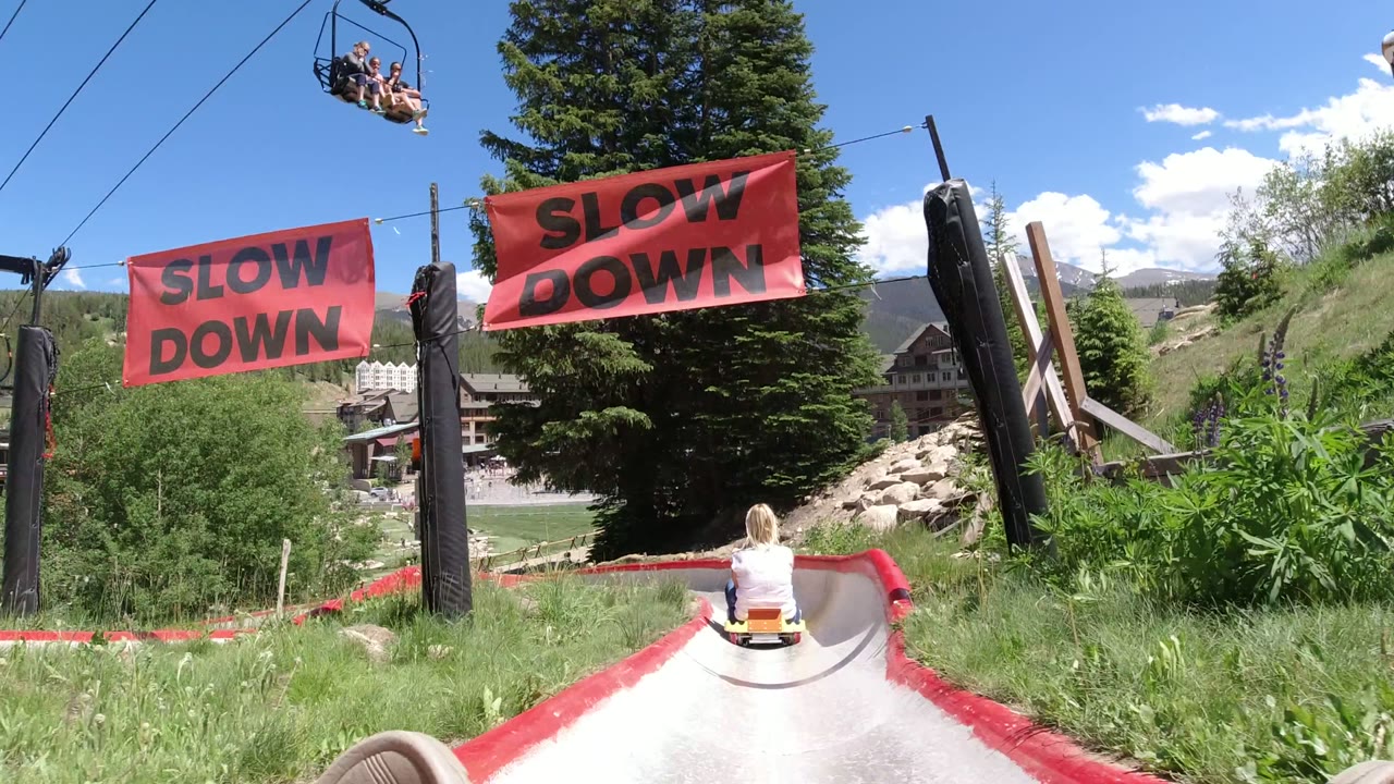 At Winter Park, Colorado, the slide passes directly under two red SLOW DOWN banners, with the rider ahead centered below chairlift cables and lodges visible beyond trees and grass.