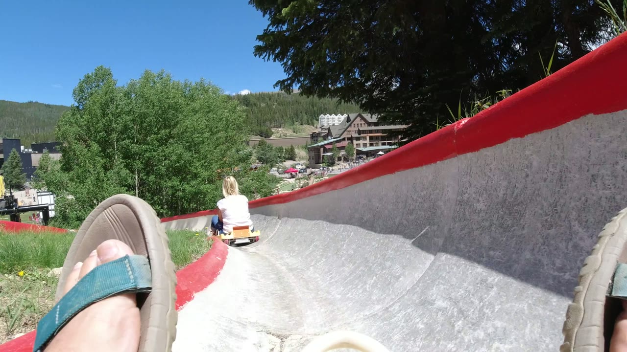 Rider-level view in Winter Park, Colorado showing a sandal in the foreground, a child ahead on a slide track, green trees, resort buildings, and mountains beyond.